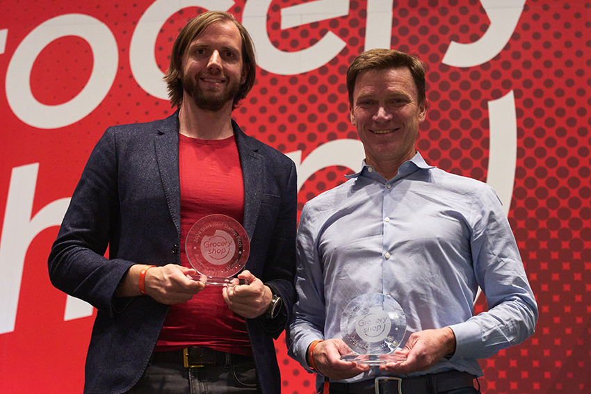 Two men holding awards in front of a red background Description automatically generated